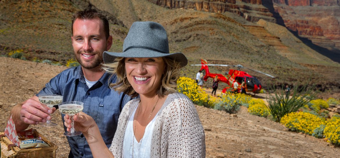 Woman and man holding champagne glasses at the bottom of Grand Canyon West with red helicopter in the background.