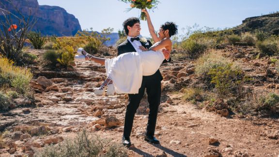 a newly married couple smile at eachother holding flowers after a helicopter landing on the bottom of the grand canyon