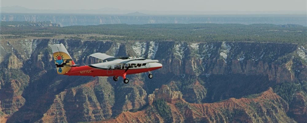 A red and white airplane flies over the Grand Canyon in a clear sky.