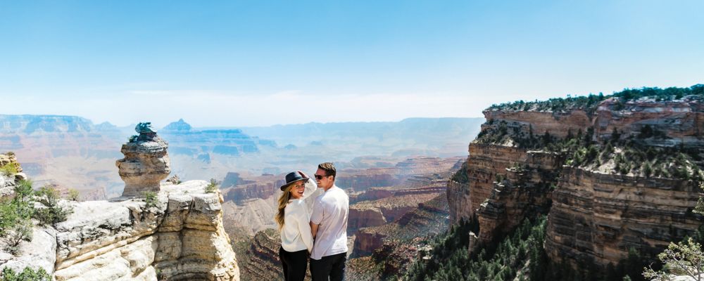 A man and woman stand at the edge of the Grand Canyon smiling and holding hands
