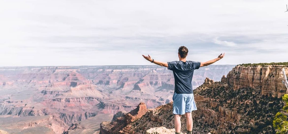 a man stands on the edge of a Grand Canyon lookout point with his arms extended in the air