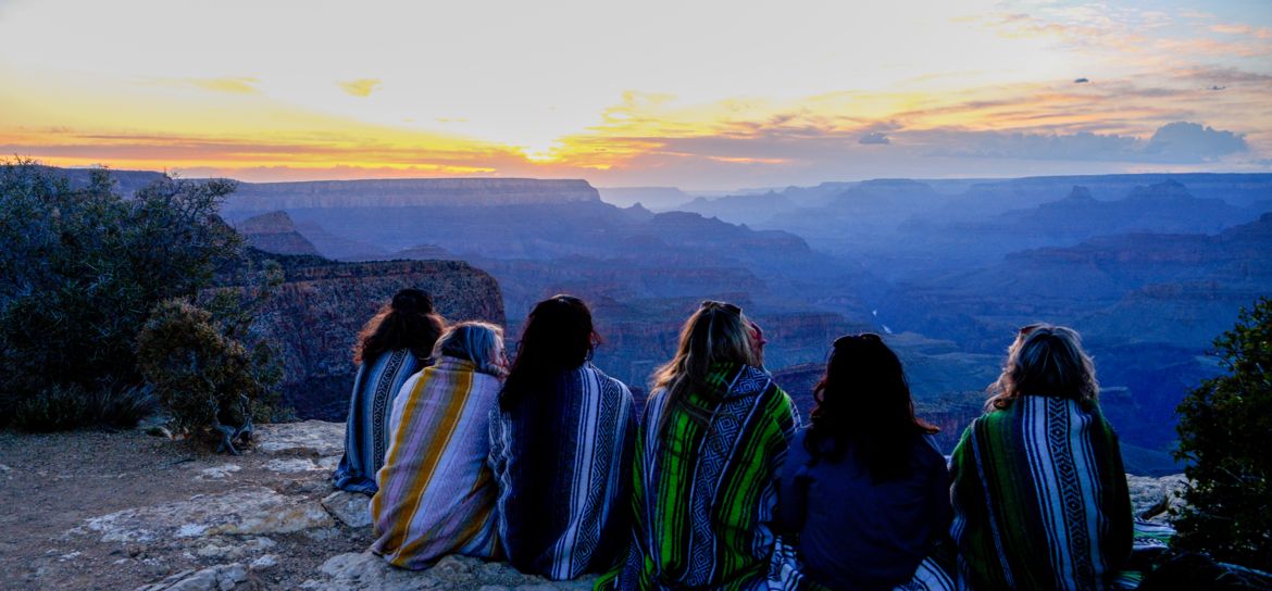a group of people sit at the edge of the Grand Canyon National Park during sunset