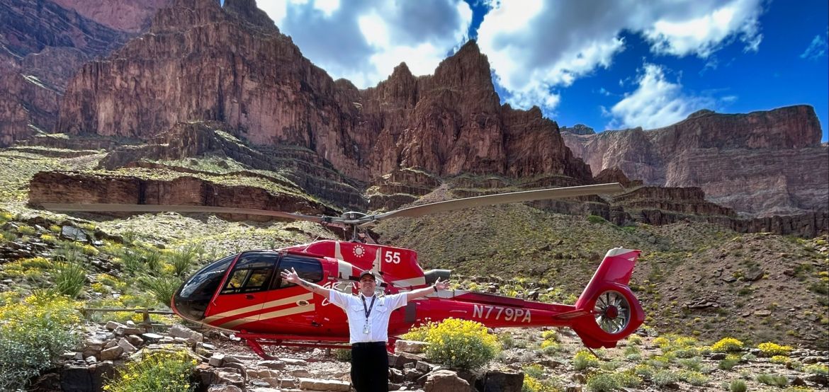 a helicopter tour pilot poses with his helicopter at the bottom of the grand canyon