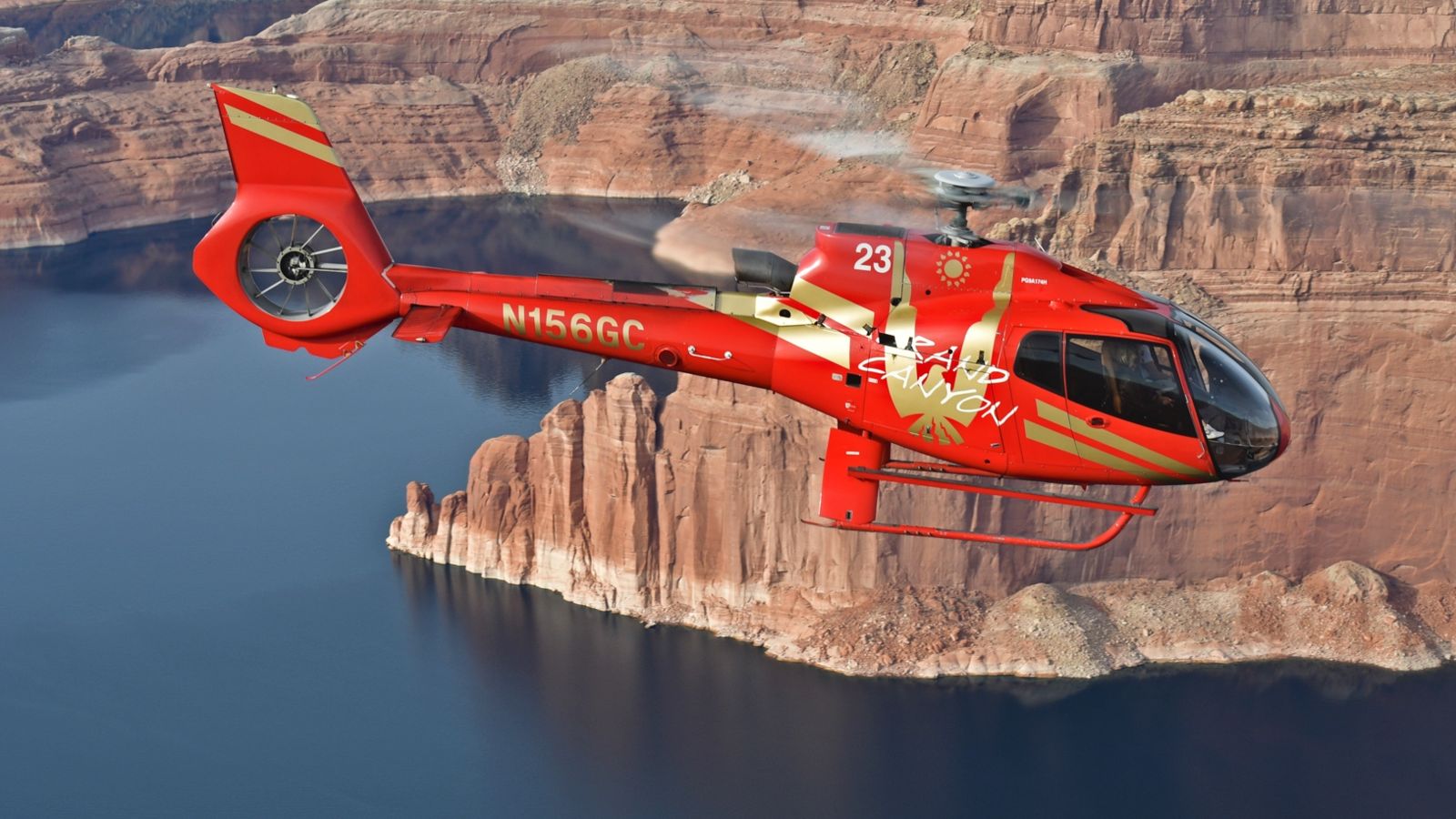 A red helicopter flies through the walls of the Grand Canyon above the Colorado River.