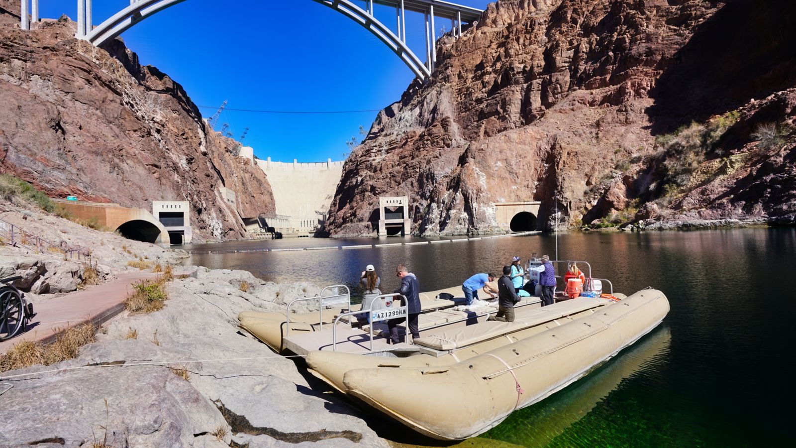 Raft carrying passengers on the Colorado River with the Hoover Dam in view from a distance.