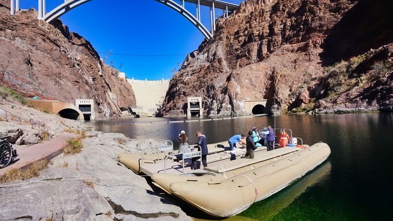 Raft carrying passengers on the Colorado River with the Hoover Dam in view from a distance.