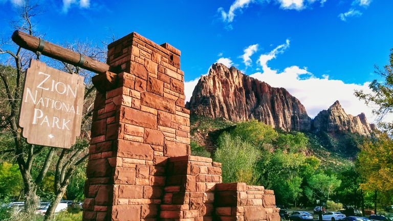 entry sign that reads “Zion National Park”, with mountains and lush forestry in the background