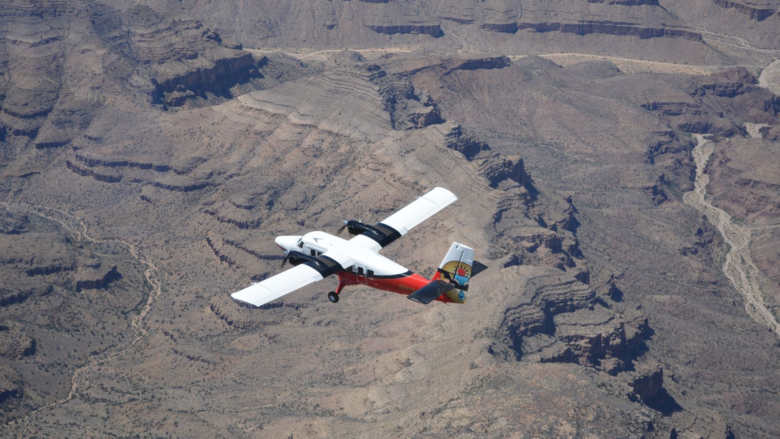 A red and white airplane mid-flight on a Grand Canyon tour, seen from above.