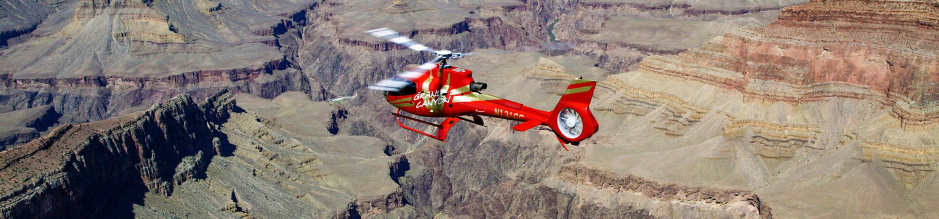 A bright red helicopter flies over the Grand Canyon and Colorado River.
