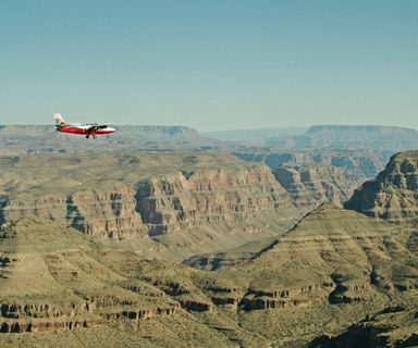 A small red and white airplane soars over a stone gorge of the Grand Canyon West Rim.