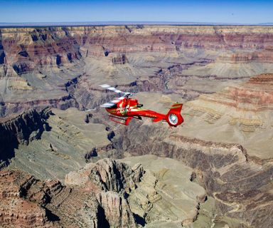 A bright red helicopter flies over the Grand Canyon and Colorado River.
