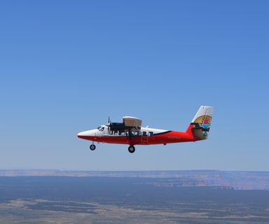 A red and white airplane flies over the Grand Canyon in a clear sky.