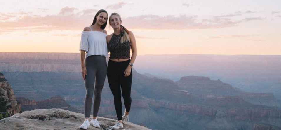 two women smile and pose at a Grand Canyon lookout point at sunset