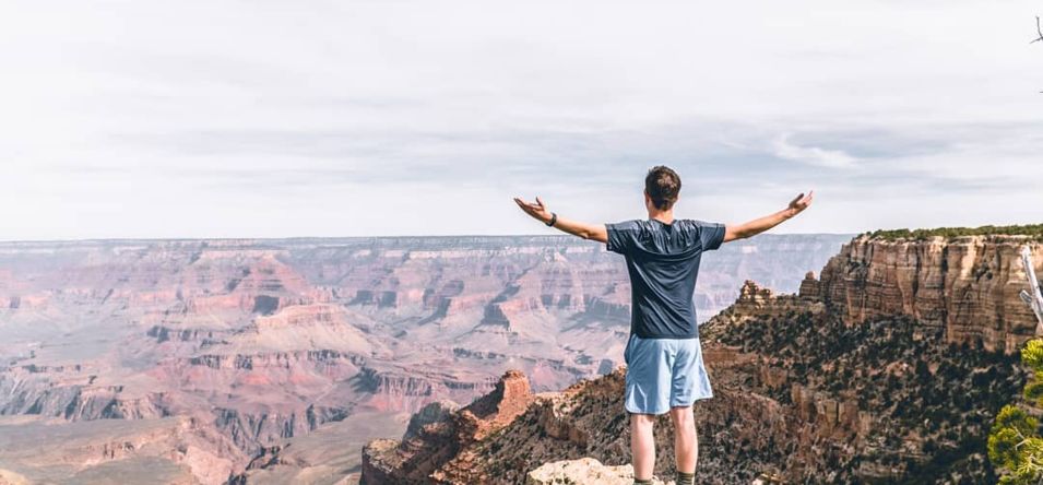 a man stands on the edge of a Grand Canyon lookout point with his arms extended in the air