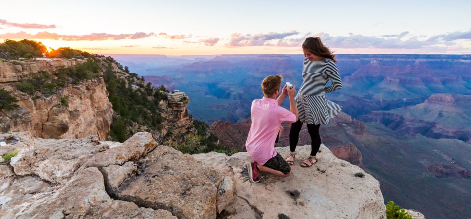 a man kneels before a smiling woman while proposing at the edge of the Grand Canyon