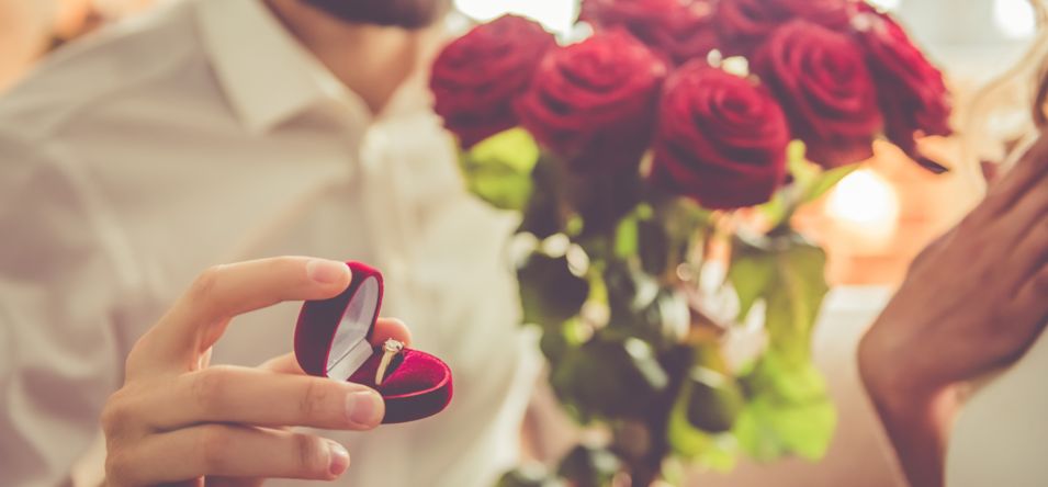 a closeup of a man kneeling before a surprised woman, holding an open ring box in one hand and a bouquet of roses in the other